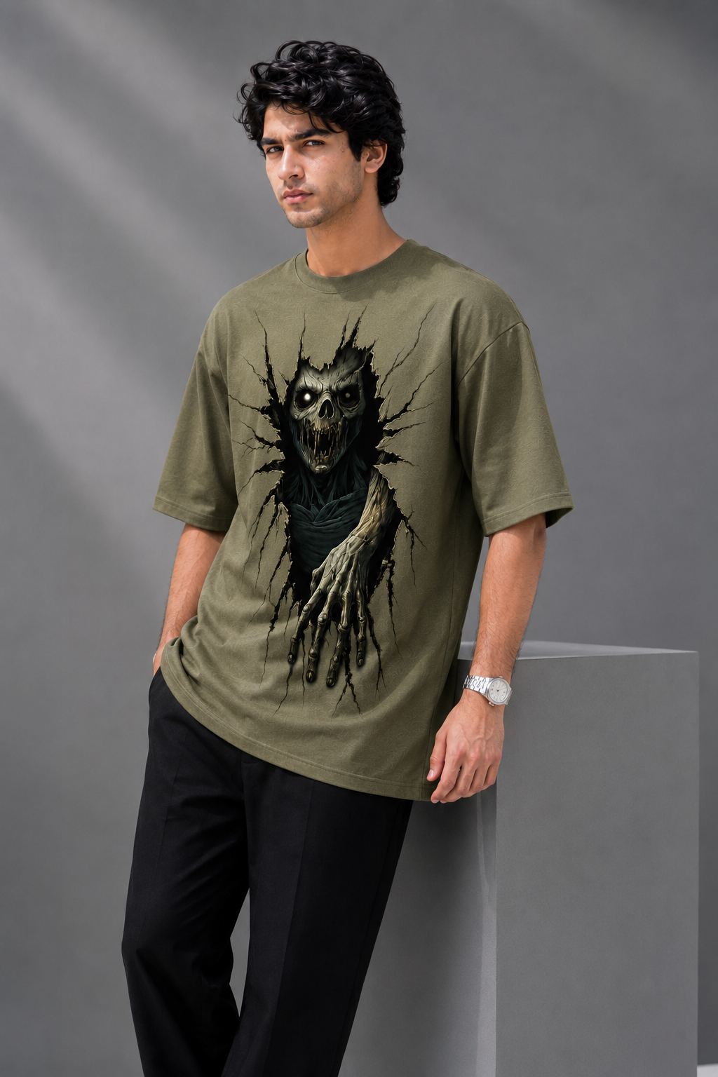 Male model wearing an AUA&CO olive green oversized baggy T-shirt with a large 3D horror skull and hand graphic print, styled with black trousers in a gray studio setting.
