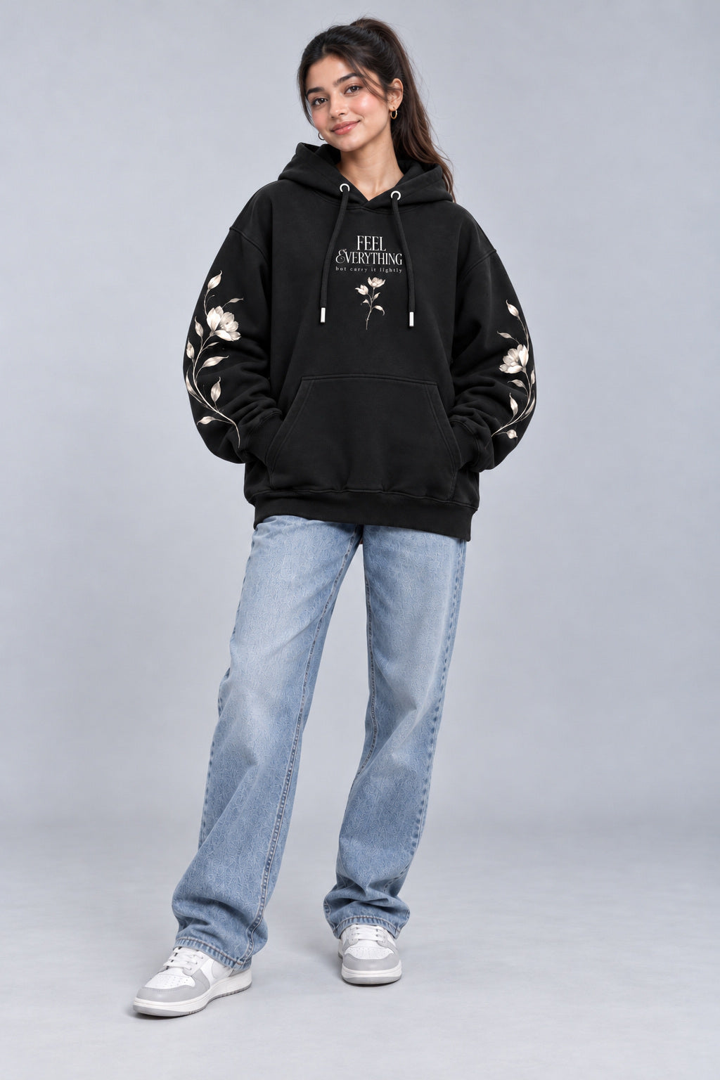 A woman modeling a black pullover hoodie with white text and floral embroidery on the sleeves, paired with light blue jeans and white sneakers, against a light gray studio background.