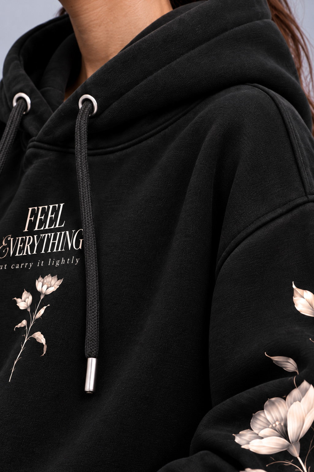Close-up of a black pullover hoodie worn by a model, featuring white text that reads 'FEEL EVERYTHING' and 'let carry it lightly' alongside white floral illustrations on the chest and sleeve.