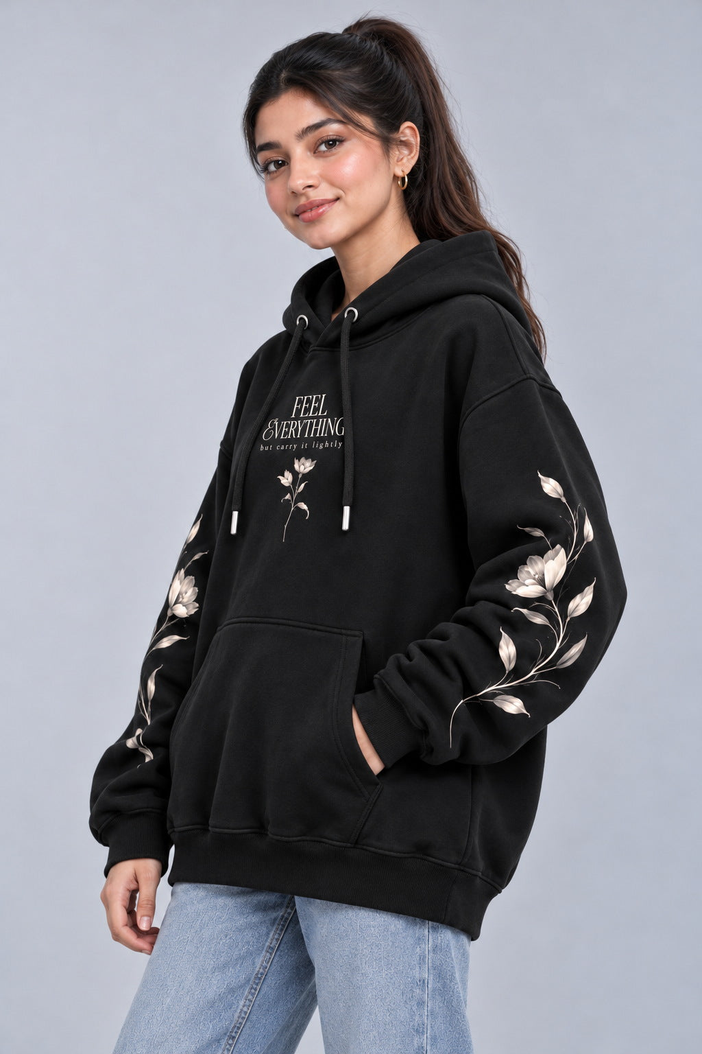 A woman modeling a black pullover hoodie with white floral embroidery on the sleeves and white text on the chest, paired with light blue jeans against a solid gray studio background.