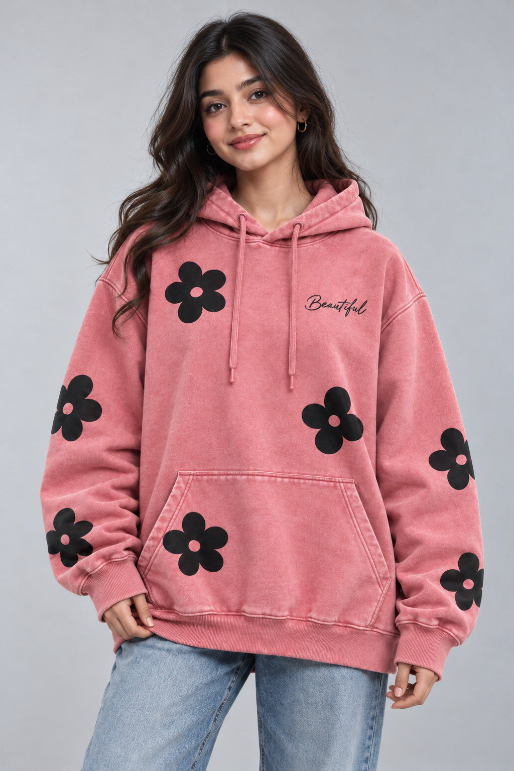 A woman wearing a pink hooded sweatshirt with black flower prints and the word 'Beautiful' embroidered on the chest, paired with light blue jeans, against a solid gray studio background.