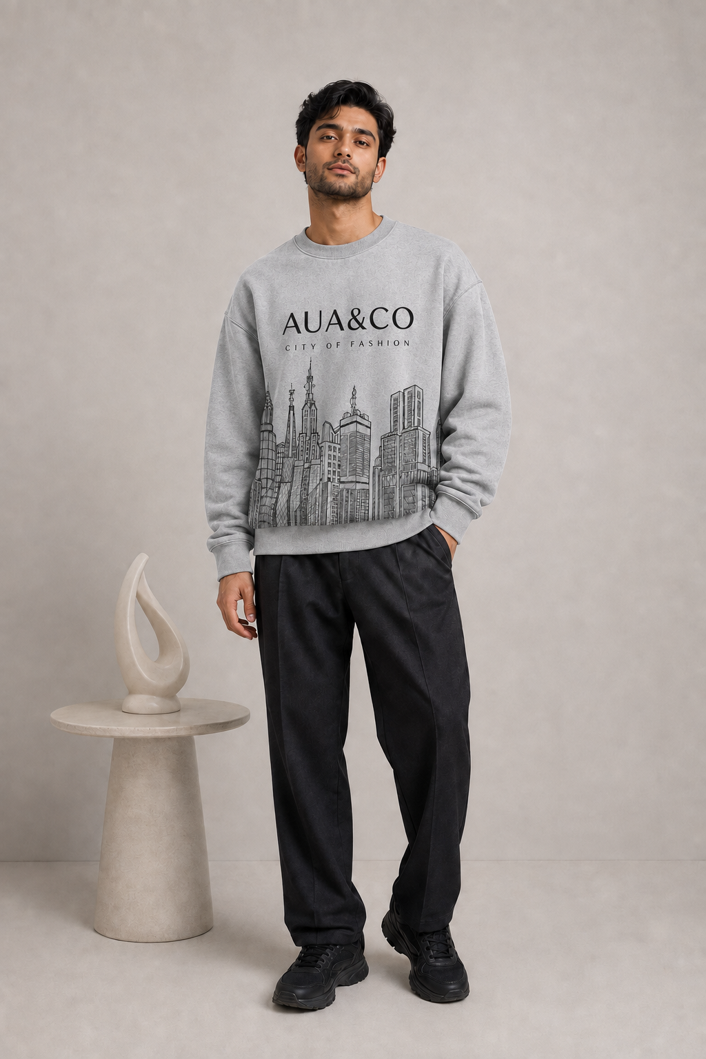 A light gray crewneck sweatshirt featuring a black line art graphic of skyscrapers and the text 'AUA&CO CITY OF FASHION' printed on the front, displayed against a neutral light gray background.