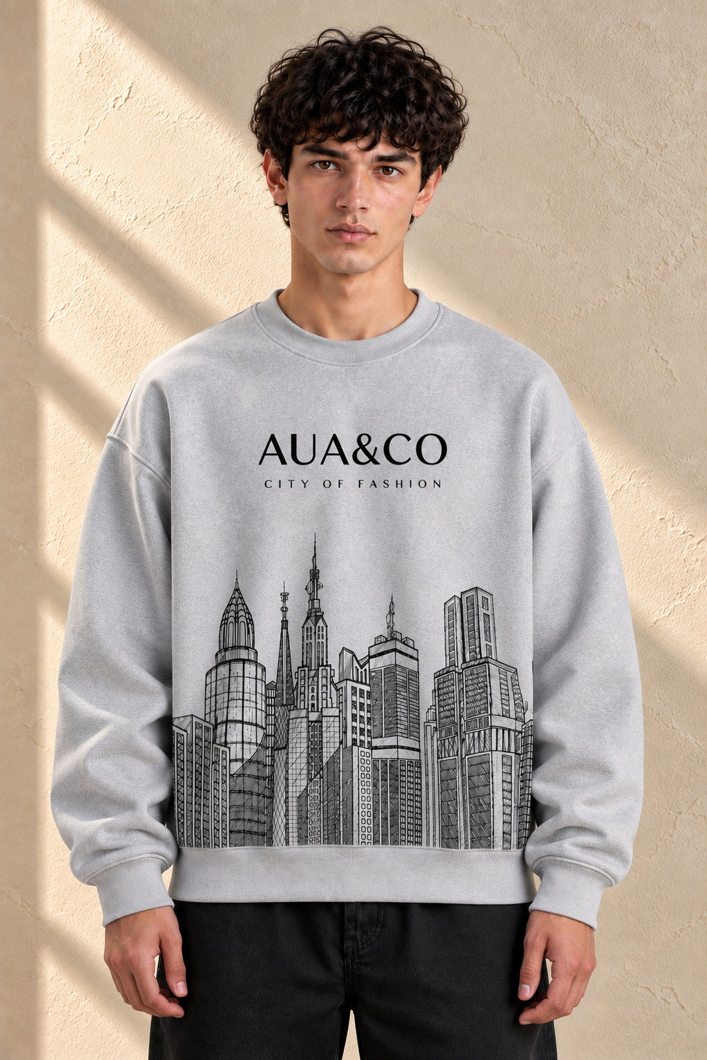 A man wearing a light gray crewneck sweatshirt with a black graphic of skyscrapers and the text 'AUA&CO CITY OF FASHION' on the front, set against a textured beige wall with soft window shadows.