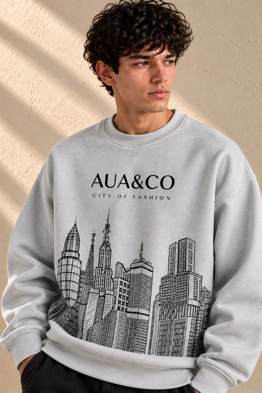 A man wearing a light gray crewneck sweatshirt with a black graphic of skyscrapers and the text 'AUA&CO CITY OF FASHION' on the front, set against a textured beige wall with soft window shadows.