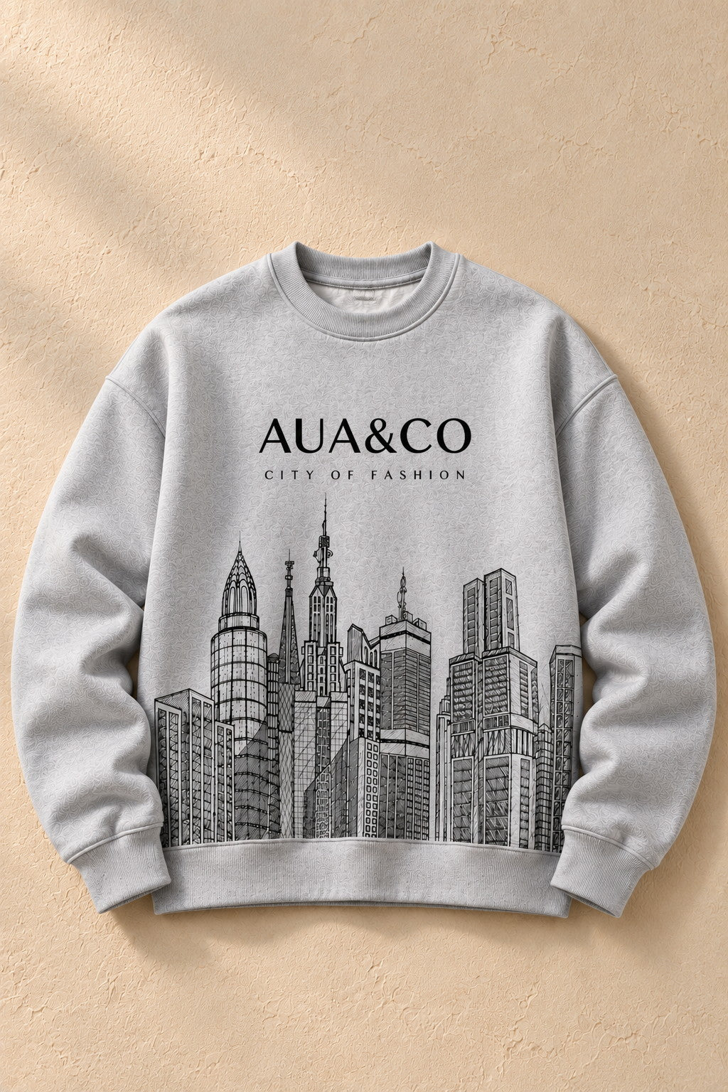 A light gray crewneck sweatshirt featuring a black graphic of a city skyline and the text 'AUA&CO' and 'CITY OF FASHION' on the front, displayed flat against a textured beige background.
