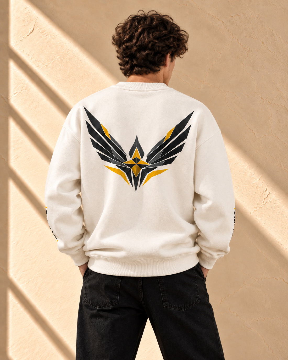 A man wearing a white crewneck sweatshirt with 'RARE BY NATURE' printed in black and yellow on the front, paired with black pants, standing against a textured beige wall with diagonal shadows.