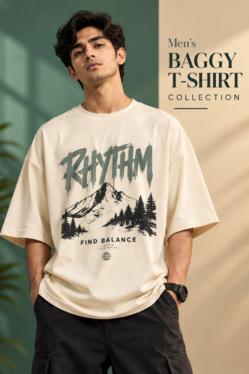 A man wearing a cream-colored oversized graphic t-shirt with a black mountain and forest print and the word 'RHYTHM' on the front, paired with black cargo pants. The background is split between a green wall on the left and a beige wall on the right with text reading 'Men's BAGGY T-SHIRT COLLECTION'.