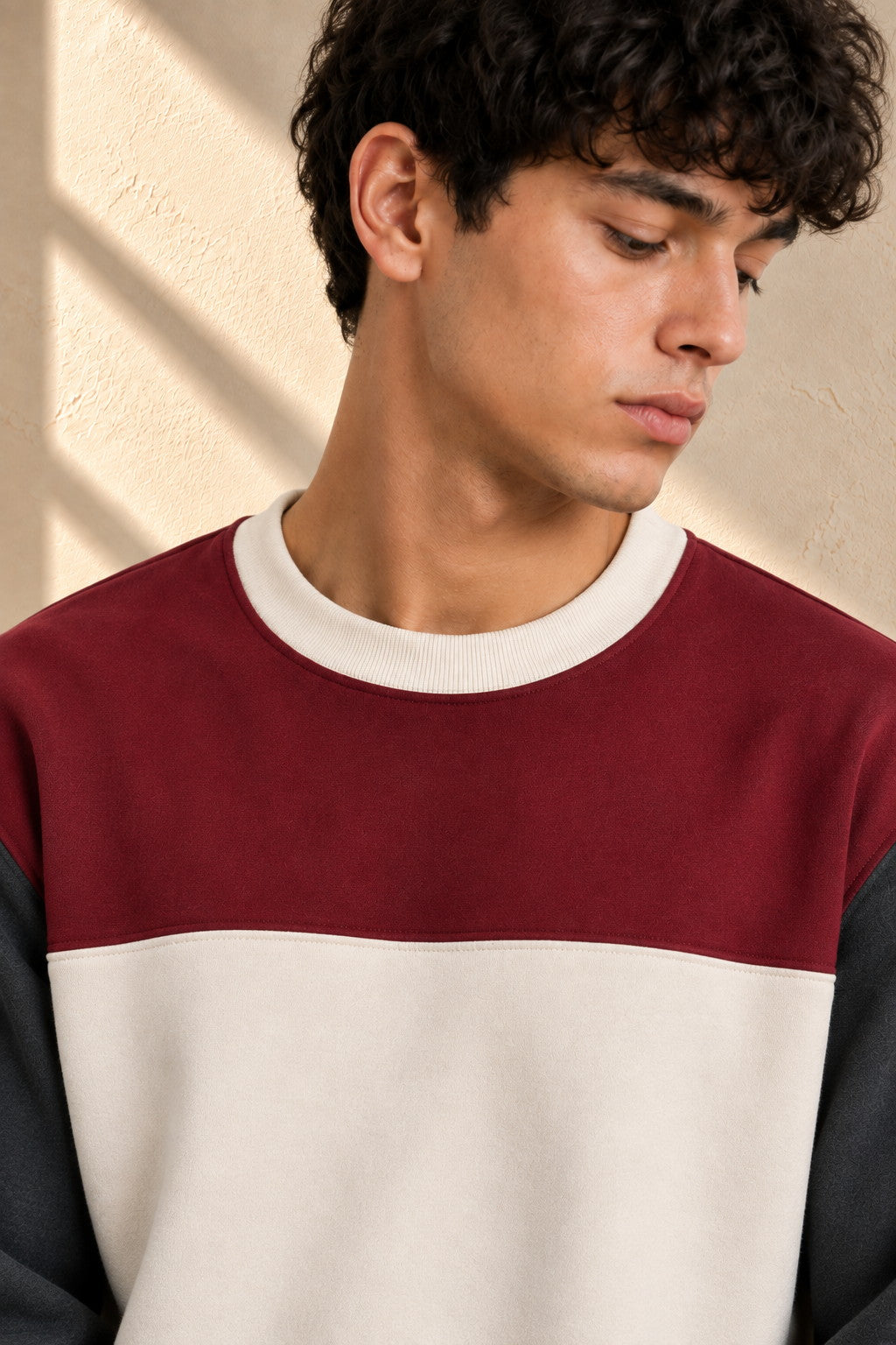 A close-up of a man wearing a colorblock crewneck sweatshirt with a burgundy upper section, a cream middle band, and dark charcoal sleeves, set against a textured beige wall with soft window shadows.