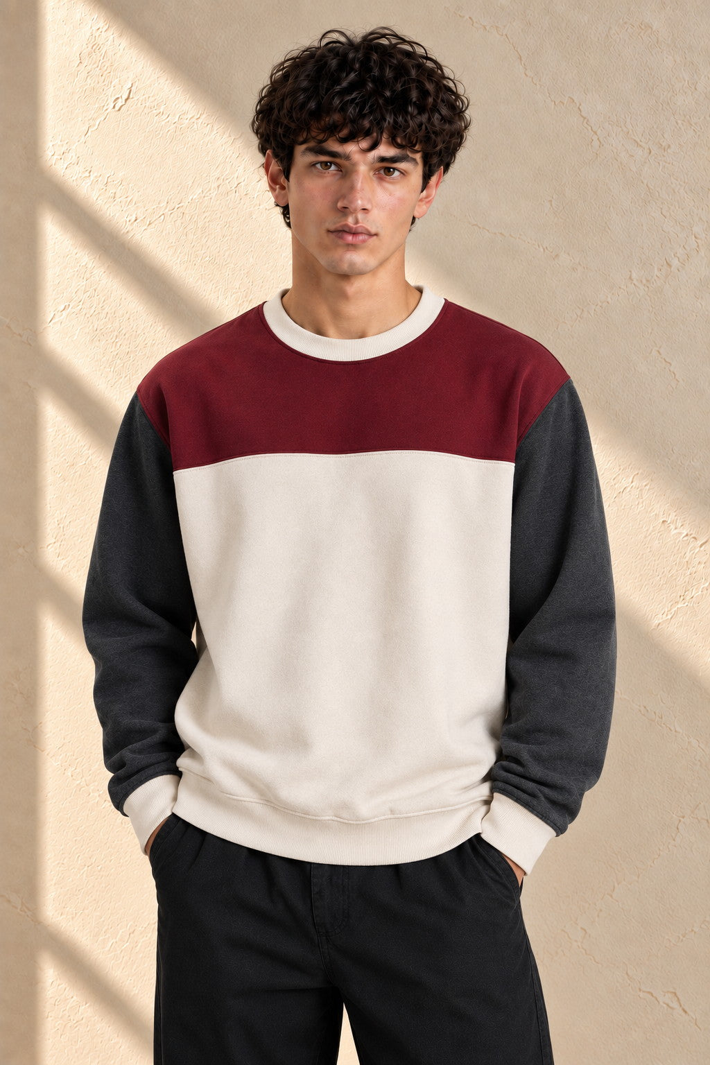 A man wearing a color-block crewneck sweatshirt with a burgundy upper chest, an off-white center, and dark gray sleeves, paired with black pants, standing against a textured beige wall with soft window shadows.
