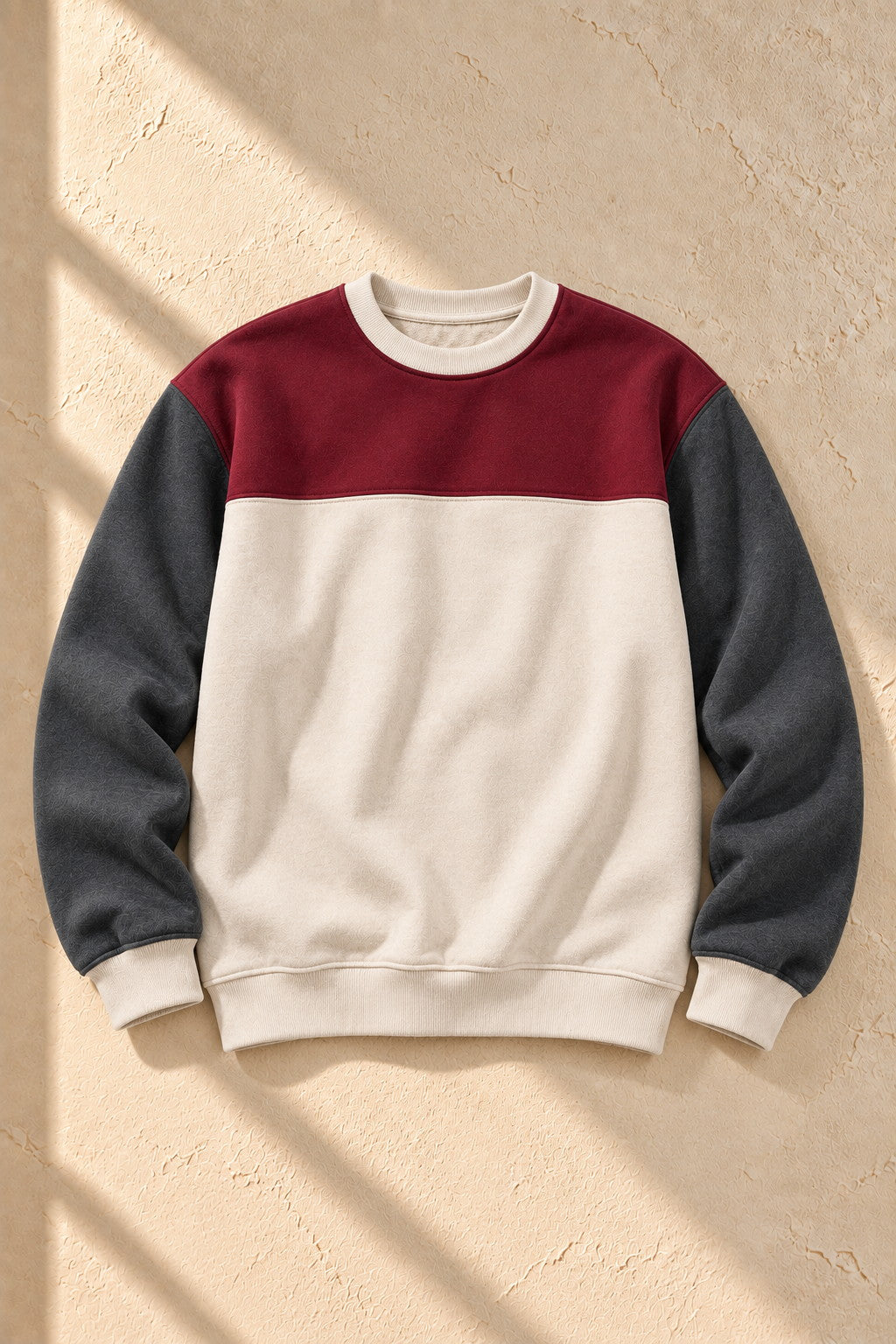 A color-block crewneck sweatshirt with a burgundy upper chest, dark gray sleeves, and an off-white body, laid flat on a textured beige wall with diagonal shadows.