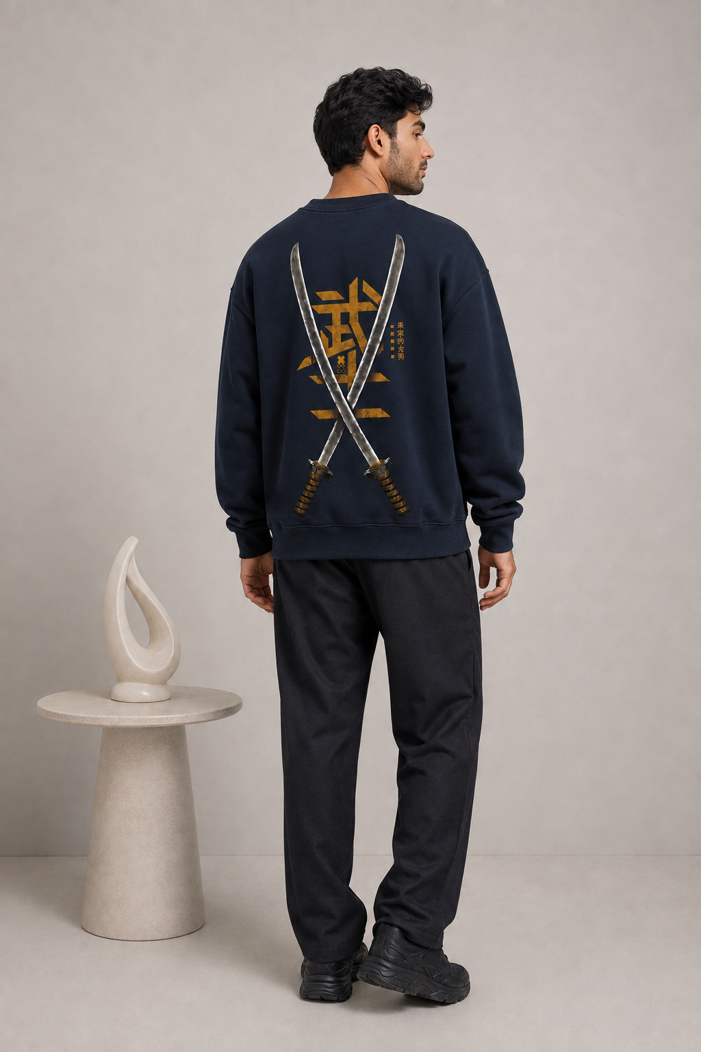 A man stands with his back to the camera wearing a navy blue sweatshirt with a large graphic of two crossed swords and gold Japanese characters on the back, paired with black pants and black sneakers, in a studio setting.