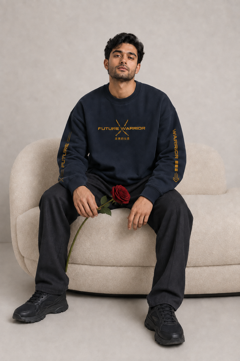 A man sits on a beige armchair wearing a navy blue crewneck sweatshirt with gold 'FUTURE WARRIOR' text on the chest and sleeves, paired with dark pants and black sneakers, holding a red rose against a neutral studio backdrop.