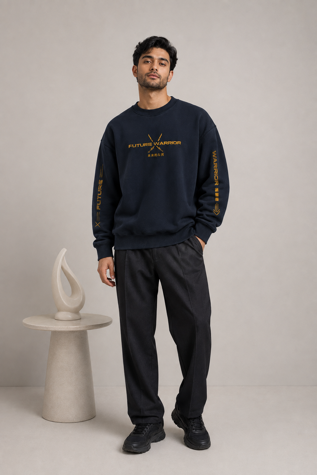 A man modeling a navy blue crewneck sweatshirt with gold 'FUTURE BARRIS PARIS' text and sleeve graphics, paired with dark gray sweatpants and black sneakers, standing in a studio next to a small sculptural side table.