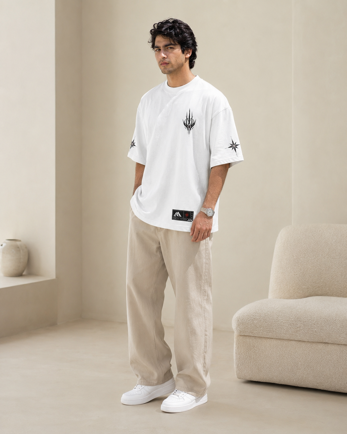 Man wearing a white t-shirt with black designs and beige pants in a minimalistic room.