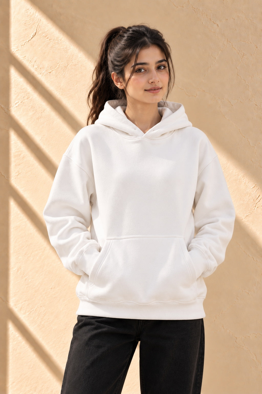 A woman wearing a plain white pullover hoodie with black pants, standing against a textured beige wall with diagonal shadows.