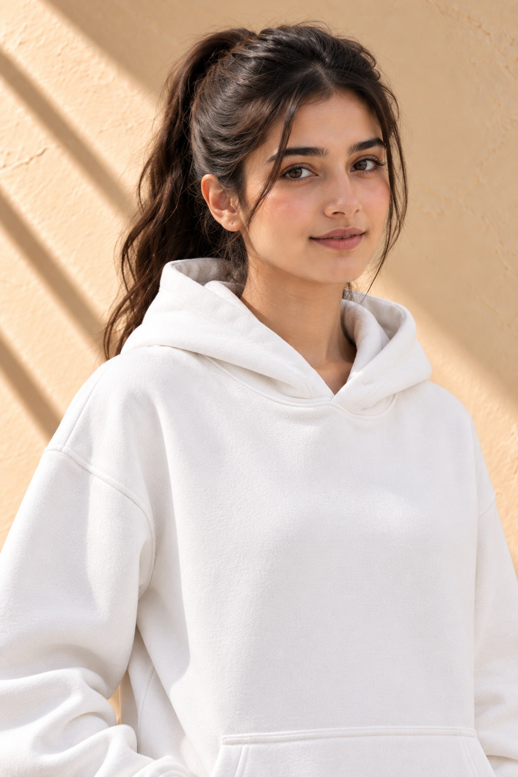 A woman wearing a plain white pullover hoodie with black pants, standing against a textured beige wall with diagonal shadows.