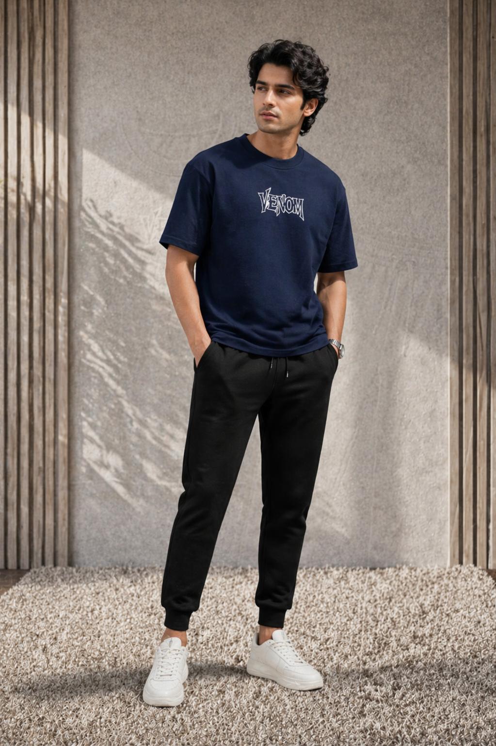 Man wearing a navy blue t-shirt with a logo and black pants standing against a textured wall.