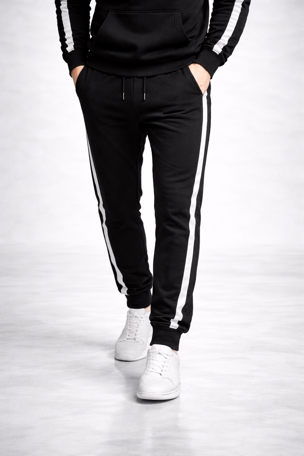 Person wearing black jogger pants with white stripes and a black hoodie on a light gray background