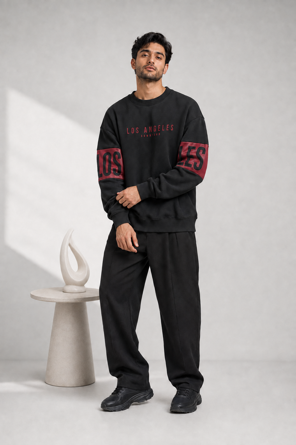 A man modeling a black crewneck sweatshirt with red and white 'Los Angeles' text on the chest and red block lettering on the sleeves, paired with black sweatpants and black sneakers, standing in a studio next to a small white decorative table.