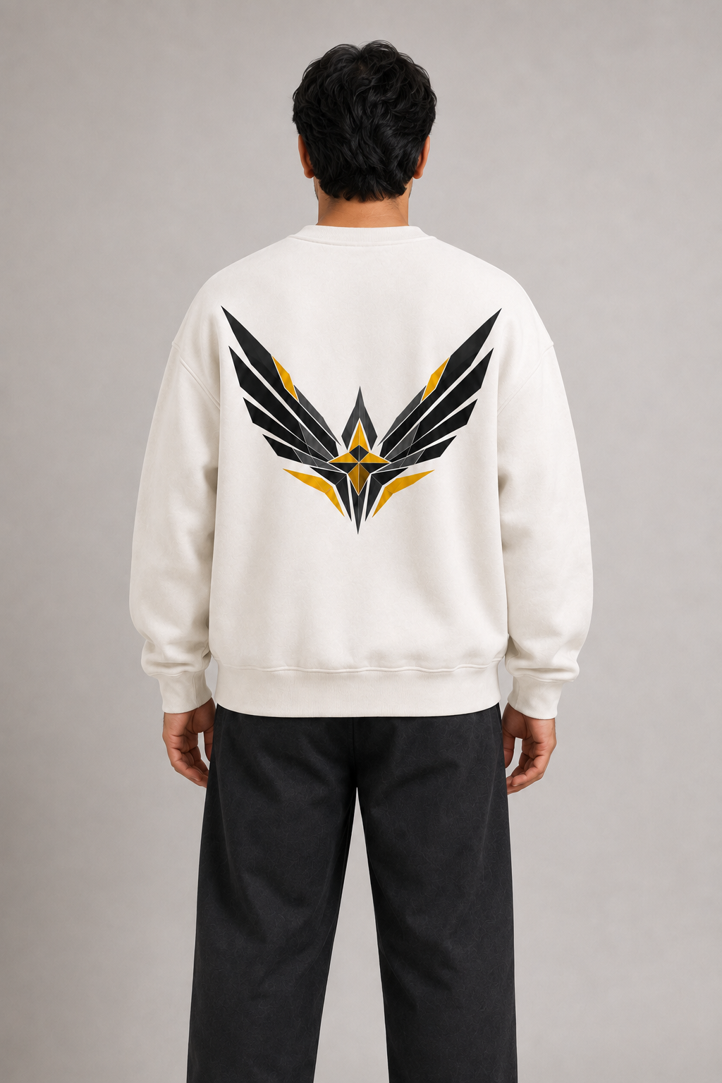 A man viewed from behind wearing a cream-colored crewneck sweatshirt with a large black and yellow winged logo printed on the back, paired with dark gray pants, against a neutral gray studio background.