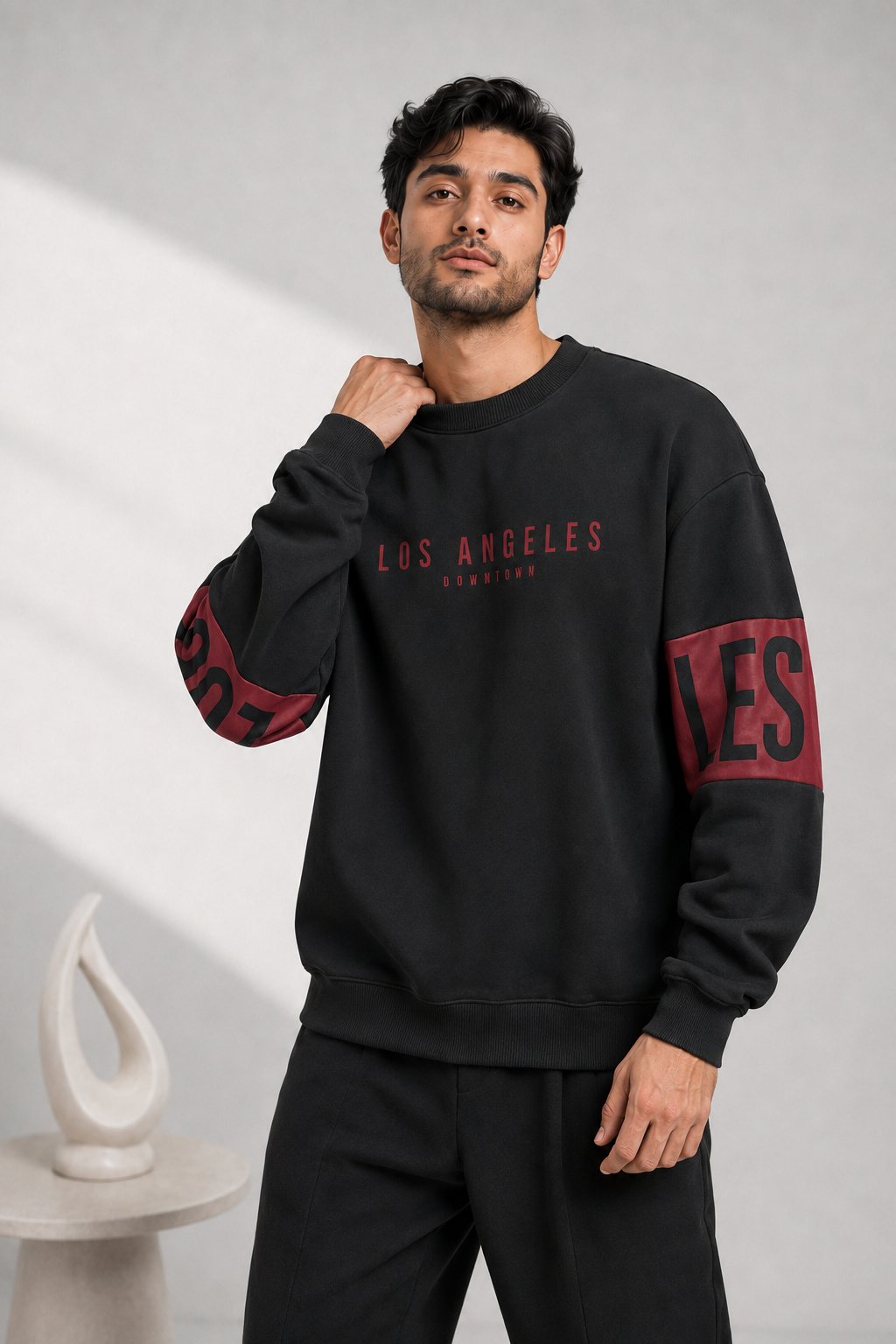 A man wearing a black crewneck sweatshirt with red and white 'Los Angeles' text on the chest and red lettering on the sleeves, paired with black sweatpants, standing against a light gray wall.