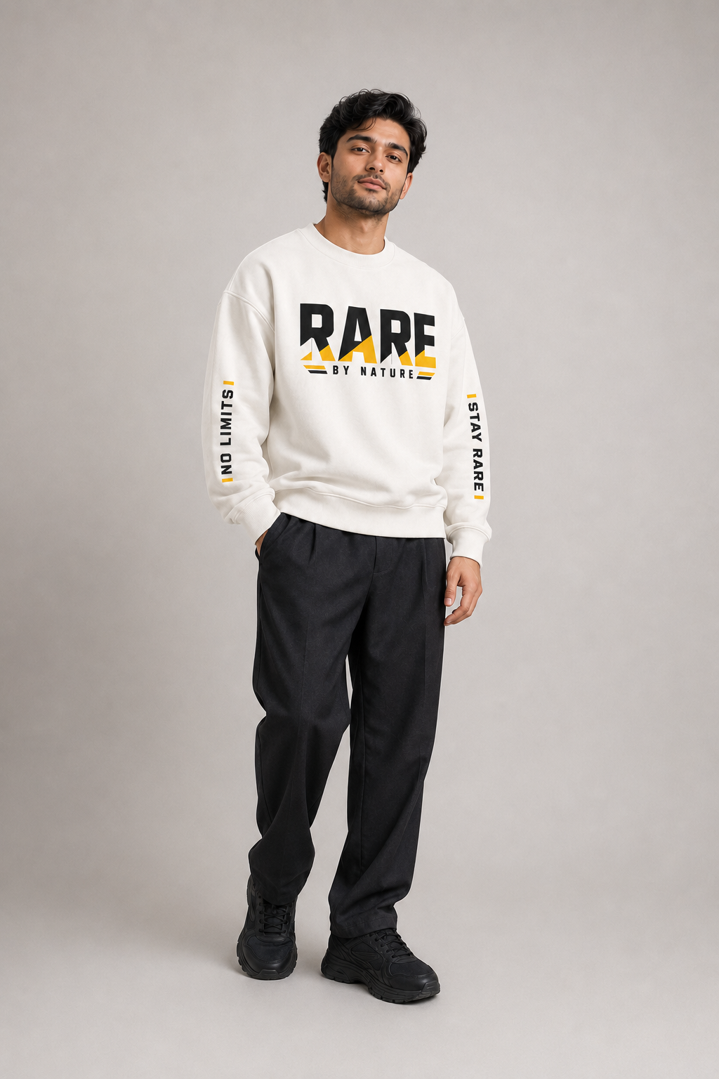 A man modeling a cream sweatshirt with bold black and yellow 'RARE BY NATURE' text on the front and sleeves, paired with black pants and black shoes, against a neutral gray studio background.